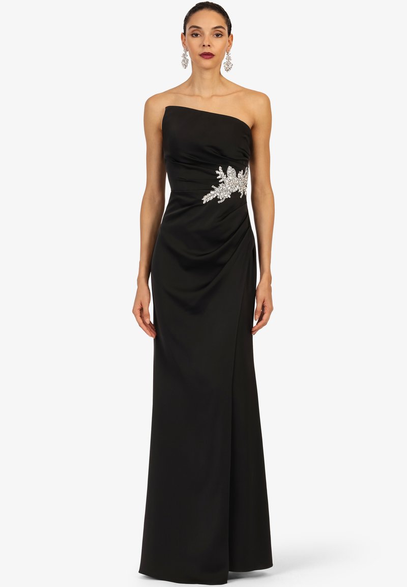 Vrouw in een strapless zwarte avondjurk met zilveren bloemenversiering op de taille, gecombineerd met hangende zilveren oorbellen.