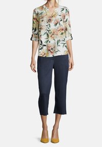 Donna che indossa una blusa a fiori con scollo a V, maniche arrotolate, pantaloni corti blu scuro e tacchi alti giallo senape, in piedi davanti a uno sfondo bianco.