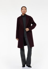 Burgundy uldfrakke med spidse slag og sidelommer, iført over en grå rullekrave og grå flagrende bukser. Sorte høje sko.