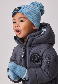 Siva puhasta bunda s kapuco, ki ima okrogel črn znak. Svetlo modra pletena kapa s cofkom in ujemajoče modre rokavice s črtami.