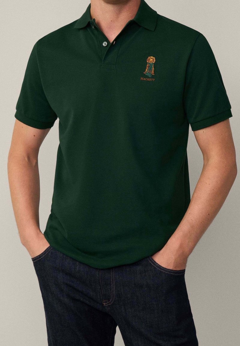 Homme portant un polo vert foncé avec un petit chien brodé et le texte « Hackett », les mains dans les poches, associé à un jean bleu foncé.
