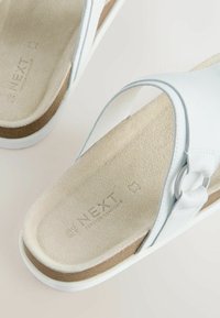 Sandales blanches à bout ouvert avec semelles intérieures beige rembourrées, brides blanches avec détail anneau argenté et semelles intermédiaires style liège.