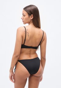 Ensemble bikini noir avec fines bretelles et finition texturée. Le bas est noué sur les côtés. Tatouage floral visible sur le haut du dos.