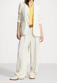 Bílá blazer s mírným polstrováním na ramenech, v kombinaci se žlutým polo tričkem a širokými, volnými bílými kalhotami. Obsahuje pletený hnědý pásek a lehké boty.