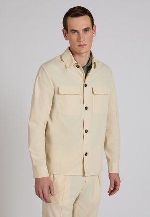 Hombre con chaqueta beige abotonada con dos bolsillos en el pecho y pantalones beige a juego, de pie frente a un fondo liso gris claro.