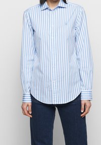 Ljusblå och vit randig skjorta med krage, långa ärmar och rundad nederkant. Har en liten logotyp på bröstet.