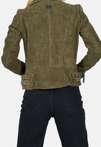 Olivgrön mockajacka med en figurnära design, synliga sömmar och två silverfärgade spännen i midjan. Kombineras med svarta jeans.