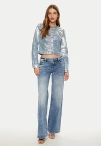 Giacca di jeans cropped blu chiaro lucida con finitura metallica e bottoni frontali, abbinata a jeans a gamba larga blu sbiadito e sandali con tacco trasparenti.