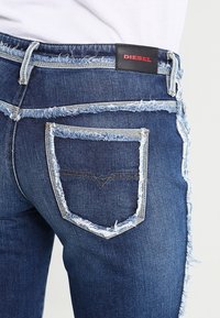 Jeans in denim blu scuro con un design consumato e bordi sfrangiati, una forma unica della tasca posteriore e un'etichetta nera con la scritta "DIESEL" in rosso.