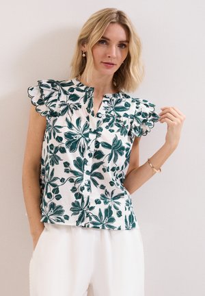 Woman with blonde hair wearing a white blouse with green floral print and ruffled sleeves, paired with white pants, against a plain background.
