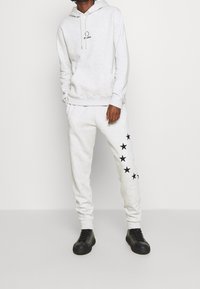 Ljusgrå hoodie med dragsko och logga, tillsammans med matchande mjukisbyxor med svarta stjärntryck på ena sidan, kombinerade med svarta sneakers.