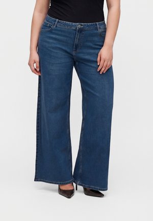 Kvinde iført højtaljede mørkeblå jeans med vide ben, kombineret med sorte spidse højhælede sko og en sort top.
