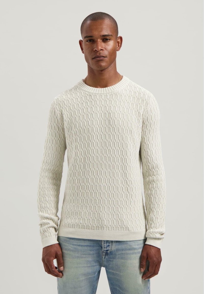 Adītas vilnas krekls gaiši krēmkrāsā ar strukturētu rakstu, apaļu izgriezumu un ribotām piedurknēm, saskaņots ar gaiši ziliem džinsiem.