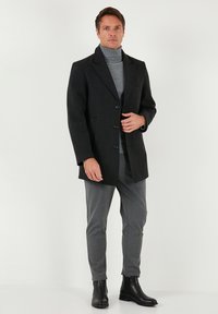 Tumehall villa segu mantel sälgkraega, kolme nupu ja kahe küljetaskuga, kantud helehalli kõrge kaelusega kampsuni ja hallide pükste peal.