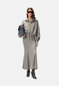 Chaqueta gris corta con cremallera y falda larga a juego, combinadas con botas negras de tacón. Cabello largo peinado en ondas, sosteniendo un bolso azul oscuro.