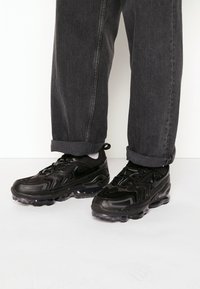 Černé tenisky Nike mají vlnitý design, síťovaný svršek a průhlednou podrážku. Nosit je s ohrnutými tmavě šedými džínami.