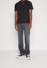 Camiseta de algodón negra con cuello redondo, que presenta un pequeño logo, combinada con jeans de mezclilla grises y zapatillas negras y blancas.
