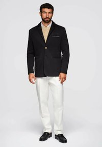 Schwarzer Wollblazer mit einem einzelnen goldenen Knopf, zwei Fronttaschen, darüber ein beiges Sweater, kombiniert mit weißen Hosen und schwarzen Schuhen.