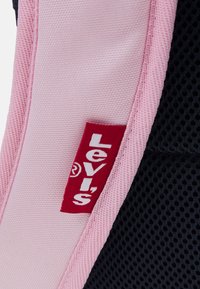 Štítek Levi's na růžovém podkladu s texturovanou síťovinou. Štítek obsahuje tučné bílé písmo na červeném obdélníkovém pozadí.