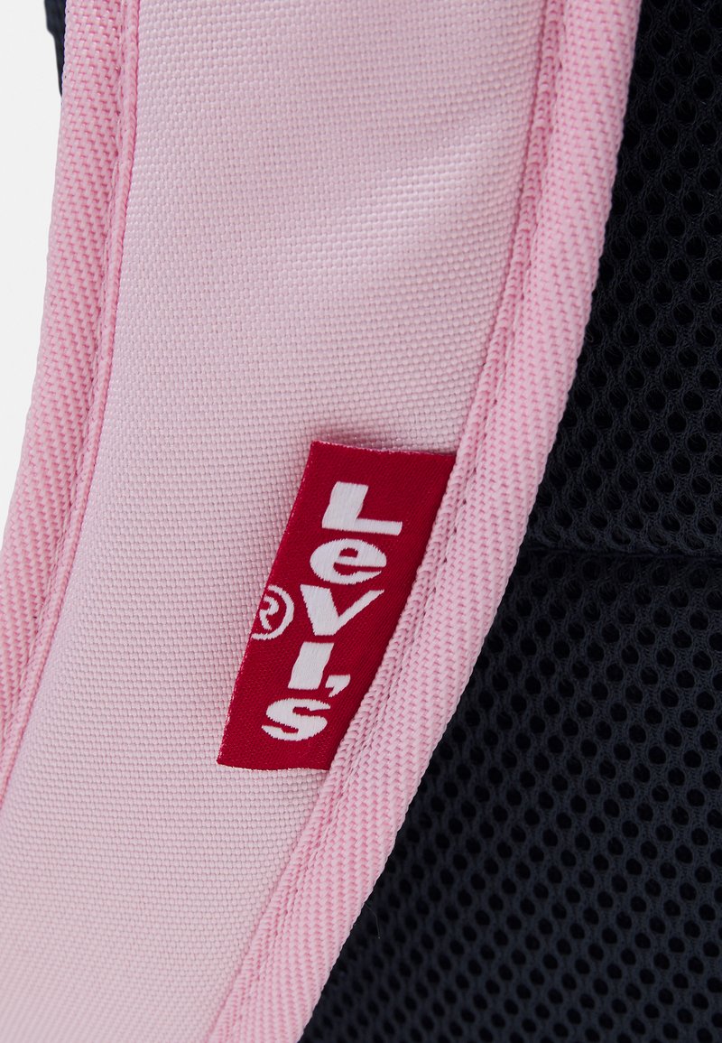 Étiquette Levi's sur un tissu rose avec un fond en maille texturée. L'étiquette présente des lettres blanches en gras sur un fond rectangulaire rouge.