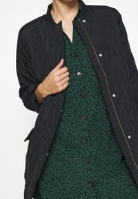 Veste matelassée noire avec col rond, fermeture éclair argentée et boutons pression portée sur une robe verte avec un motif floral noir.
