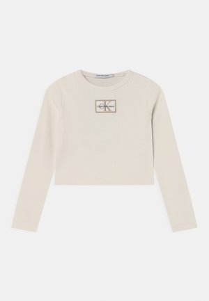 Jasno beżowy krótki top z długim rękawem, wykonany z prążkowanej tkaniny, z prostokątną naszywką z logo "Calvin Klein Jeans" oraz akcentami "K".