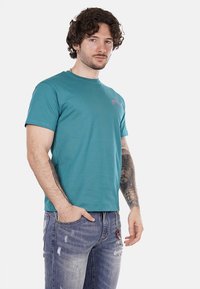 T-shirt corto color teal con scollo rotondo. Presenta un logo sottile sul lato sinistro del petto e una texture morbida e liscia. Indossato con jeans strappati.