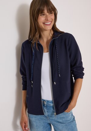 Femme souriante portant une veste zippée bleu marine avec cordons sur un haut blanc et un jean bleu clair, debout contre un mur clair uni.