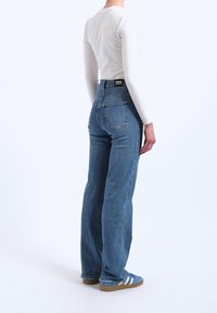 Personne portant un jean droit bleu, un haut blanc à manches longues et des baskets bleues avec des rayures blanches, debout devant un fond uni.