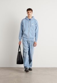 Lichtblauwe zip-up hoodie met voorkruiszakken, gecombineerd met lichtdenim jeans en zwarte instapschoenen. Een zwarte tote bag wordt in de linkerhand vastgehalten.