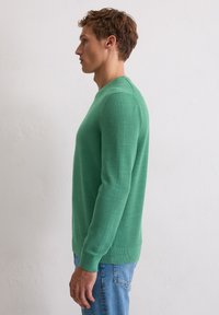 Grüner Strickpullover mit strukturierter Oberfläche, rundem Ausschnitt und langen Ärmeln. Die Passform ist schmal und wird mit blauen Jeans kombiniert.