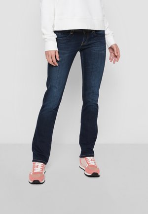 Jean en denim bleu foncé avec une coupe droite, présentant des poches avant et une fermeture éclair, associé à des baskets roses et un sweatshirt blanc.