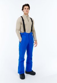 Blauwe sneeuwbroek met verstelbare zwarte bretels, voorzien van zijzakken en een rechte pasvorm. Gedoopt met een beige longsleeve top en zwarte laarzen.