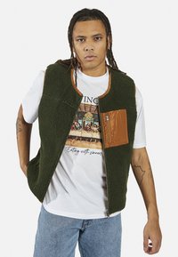 Colete de polar verde com detalhes em nylon castanho e um bolso com fecho. Usado sobre uma t-shirt gráfica branca e combinado com jeans azul claro.