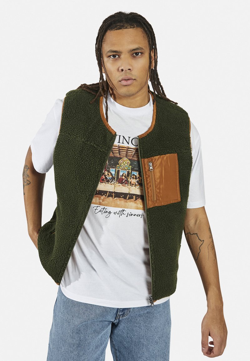 Colete de polar verde com detalhes em nylon castanho e um bolso com fecho. Usado sobre uma t-shirt gráfica branca e combinado com jeans azul claro.
