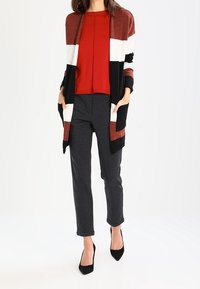 Femme portant un chemisier rouge, un cardigan bicolore noir et rouille, un pantalon gris et des escarpins noirs à bouts pointus, debout les mains dans les poches.