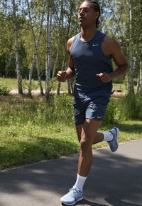 Jogador atlético en una camiseta sin mangas azul marino y pantalones cortos a juego, usando zapatillas de correr azul claro con detalles en blanco. Entorno al aire libre con árboles.