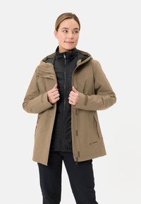 Giacca esterna beige con cappuccio, dotata di una zip anteriore, due tasche frontali e fodera interna nera. Indossata sopra una giacca intermedia nera.