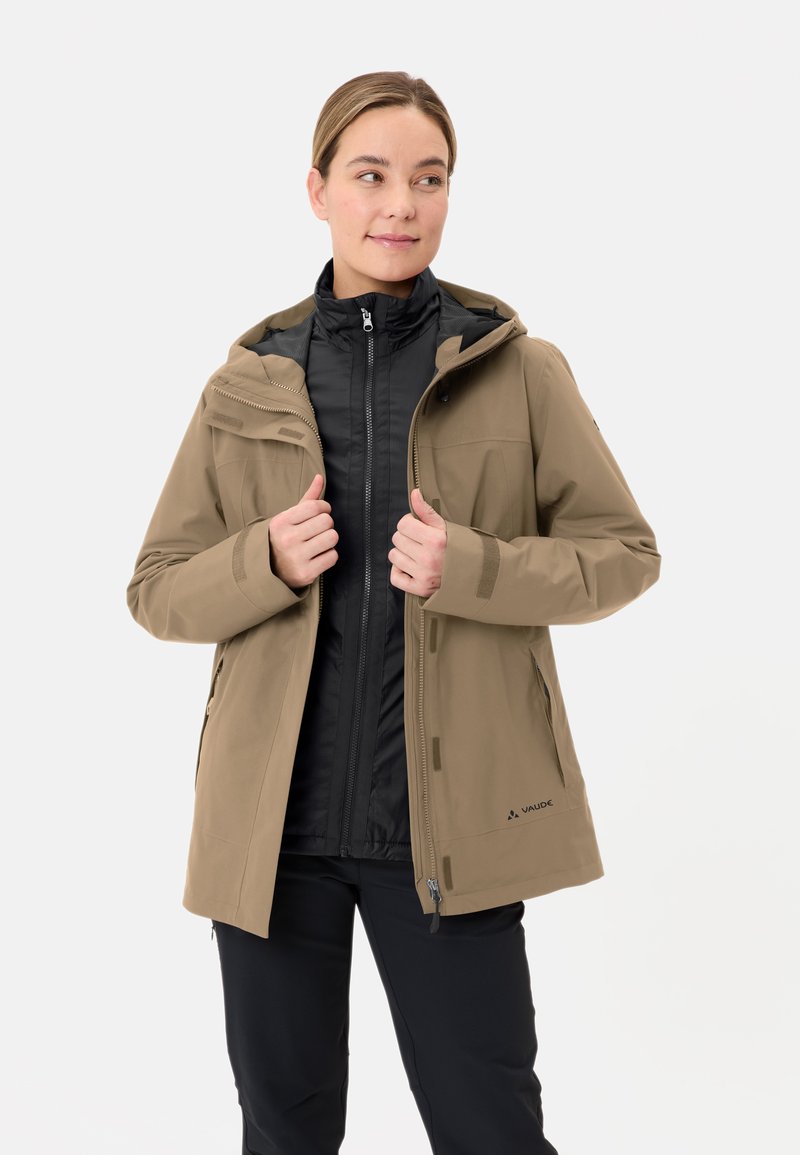 Giacca esterna beige con cappuccio, dotata di una zip anteriore, due tasche frontali e fodera interna nera. Indossata sopra una giacca intermedia nera.