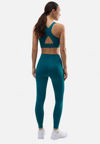 Sujetador deportivo teal con diseño de espalda cruzada, que presenta un detalle recortado. Combinado con leggings high-waist teal y zapatillas deportivas blancas.