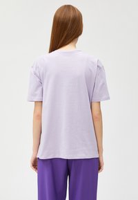 Camiseta de algodón lila con un corte relajado, mangas cortas y cuello redondo, vista desde la parte de atrás. Textura suave, costuras mínimas.