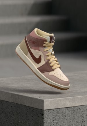 Pantof sport Nike Air Jordan cu talpă înaltă în tonuri bej și maro, șireturi legate și suspendat deasupra unei suprafețe din beton cu scări în fundal.