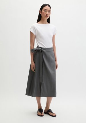 Frau trägt ein weißes kurzärmliges Oberteil, einen grauen Midi-Wickelrock, der in der Taille gebunden ist, und schwarze offene flache Sandalen, steht vor einem weißen Hintergrund.