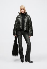 Jachetă matlasată din piele neagră, design scurt, cu fermoare, asortată cu un pulover cu guler înalt negru și blugi evazați închiși la culoare, ținând o geantă de blană neagră.