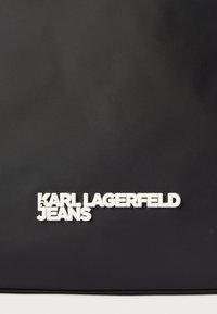 Μαύρη νάιλον τσάντα με ανάγλυφο, λευκό λογότυπο «KARL LAGERFELD JEANS» στο κάτω μέρος. Λείο υλικό χωρίς επιπλέον σχέδια ή διακοσμητικά στοιχεία.
