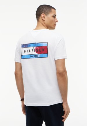 Junger Mann trägt ein weißes T-Shirt mit einem blauen, weißen und roten Grafikblock und Text "HILFIGER Est. 1985" auf dem Rücken.