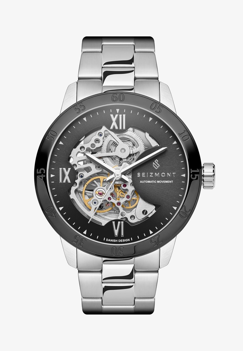 Herreklokke med gjennomsiktig mekanisk urverk; armbånd i rustfritt stål; svart skive med romerske tall; sølvaksenter og gullgir.