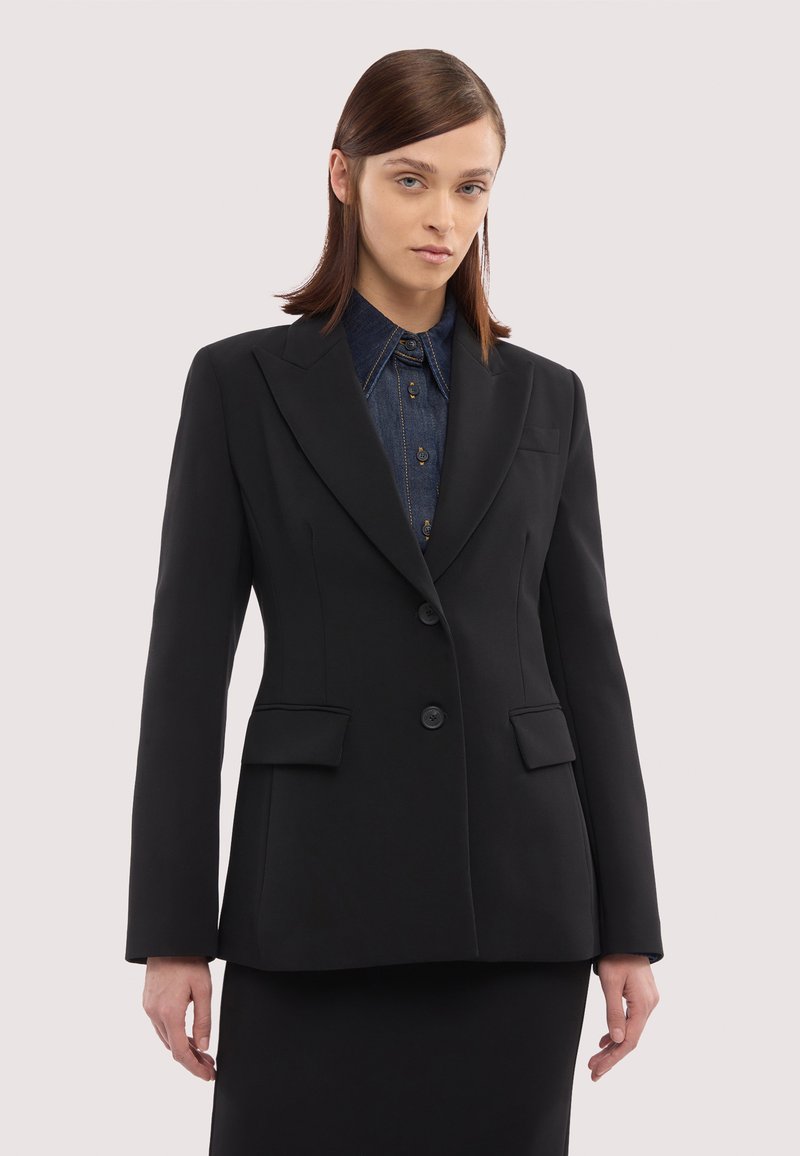 Kvinde med glat brunt hår iført en tætsiddende sort blazer over en mørk denimskjorte med knapper mod en ensartet lys baggrund.