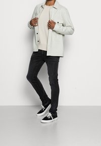 Man adjusting light grey button-up jacket, wearing beige sweatshirt, black skinny jeans, and black sneakers with white soles and laces.