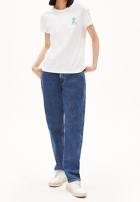 Camiseta blanca de algodón con un logo gráfico verde, combinada con jeans de mezclilla azul de pierna recta y zapatillas blancas con suelas beige.